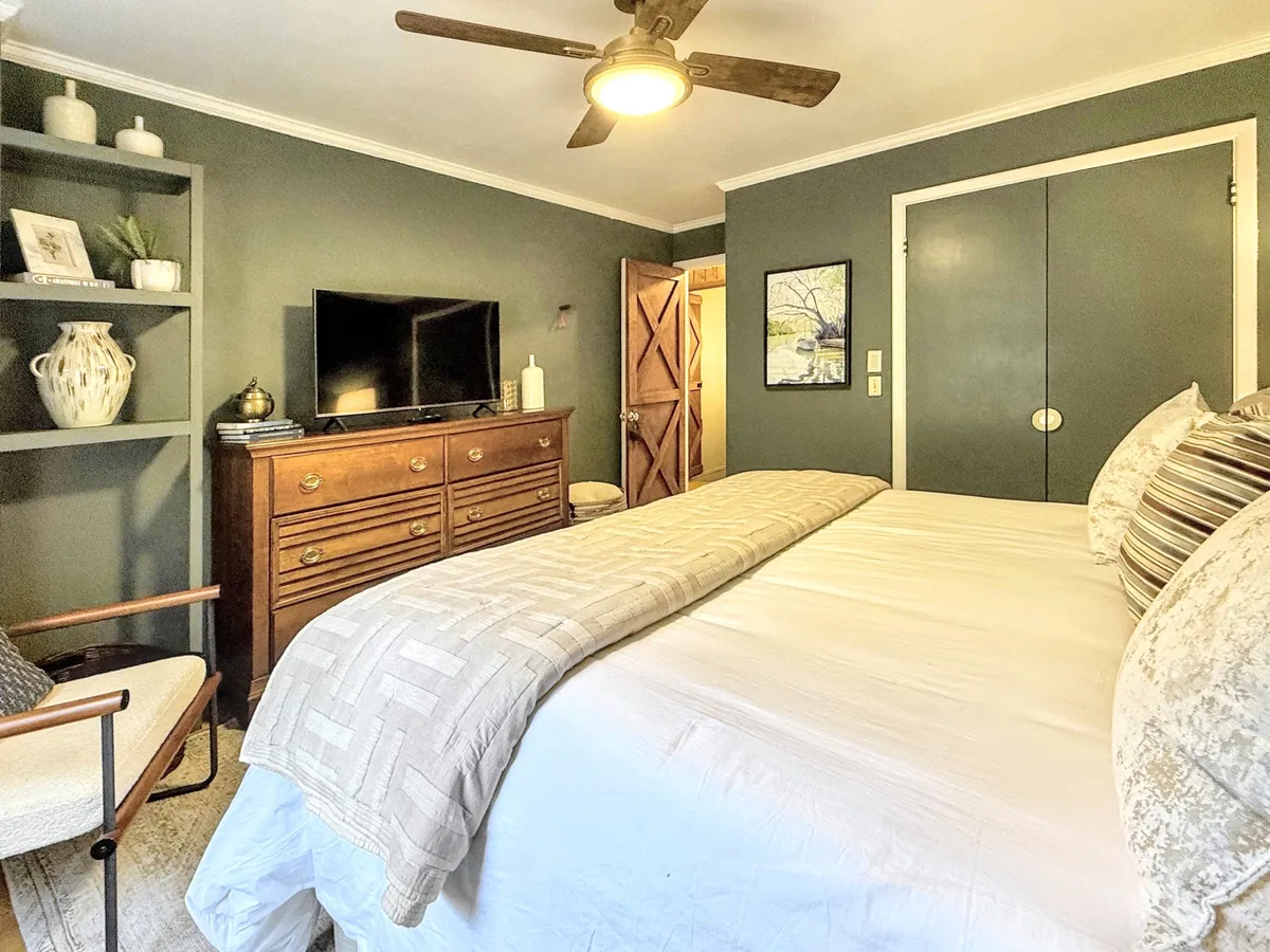 A well-appointed bedroom featuring a bed with a cozy comforter, a dresser, shelving unit, and a television. Artwork on the wall and natural light contribute to a comforting, versatile atmosphere.