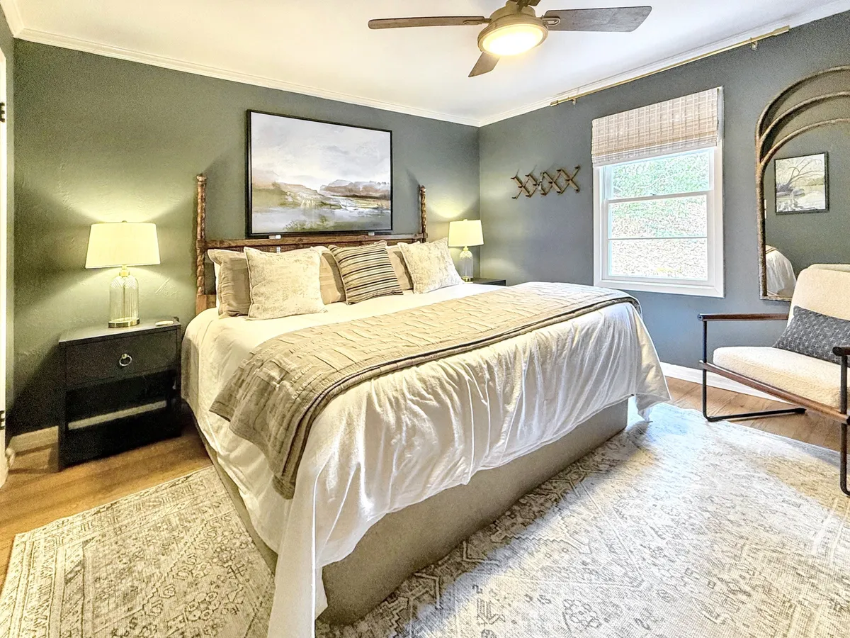 A spacious king master suite featuring a large bed adorned with decorative pillows, landscape artwork above the headboard, side tables with lamps, and a large window that bathes the room in natural light. The restful setting invites relaxation and quiet moments.
