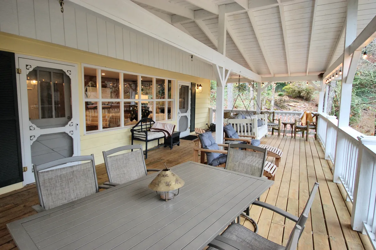 A view of the covered deck with a dining table, swing bench, and comfortable outdoor seating, complemented by large windows that connect the space to the outdoors. The area appears ready for socializing, dining, or relaxing, with natural light and greenery visible.