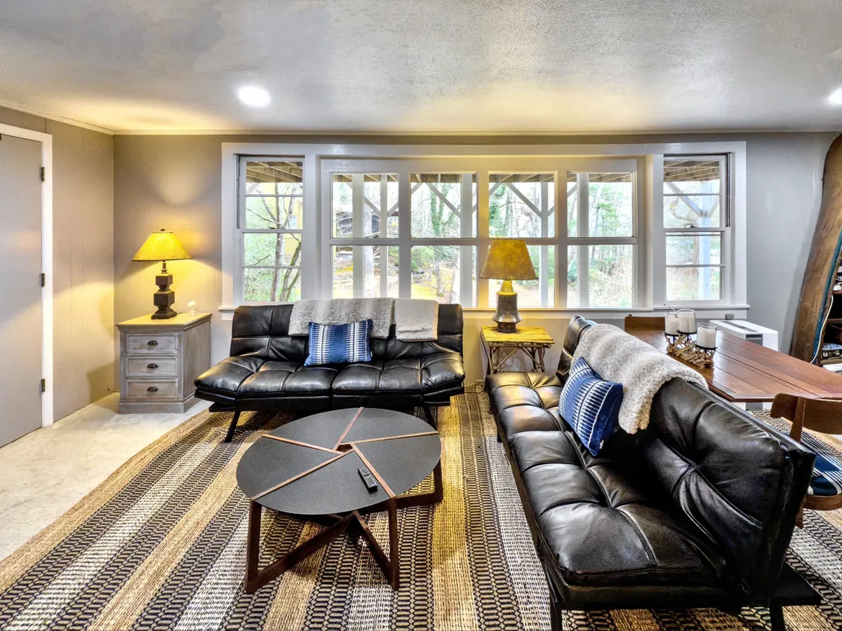 A bright, open seating area with black leather sofas, a round coffee table, a warm lamp, and large windows that offer a view outside. The arrangement encourages socializing, relaxing, and enjoying natural light, all crucial for group stays.