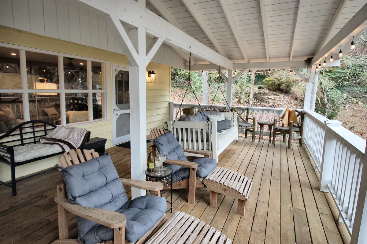 A calm and cozy exterior shot of a wooden deck featuring rocking chairs, a swing seat, and a decorative throw blanket. Wine glasses sit on the table, suggesting a space that blends leisure and work, surrounded by the tranquil outdoors.