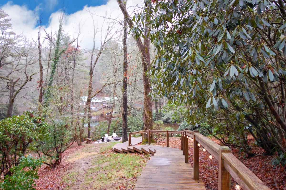 A serene exterior pathway made of wood, leading through lush trees with a view of water in the distance. The environment is tranquil, ideal for nature walks or taking a break from work for relaxation and reflection.