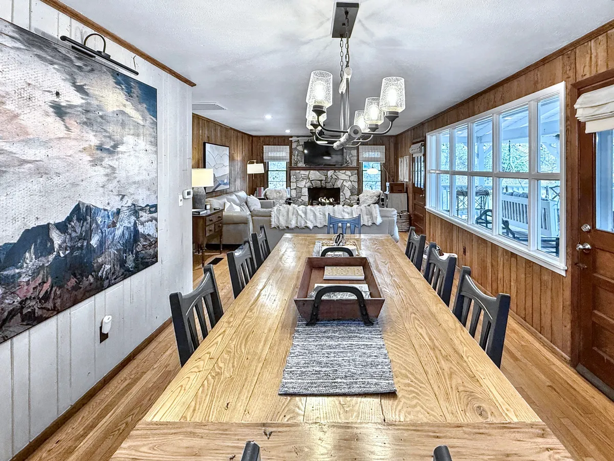 A long dining table sits beneath a chandelier, with artwork on the wall and a view into a cozy living area featuring a fireplace. The open, communal setting suggests an ideal environment for large family meals, group celebrations, or friendly gatherings.