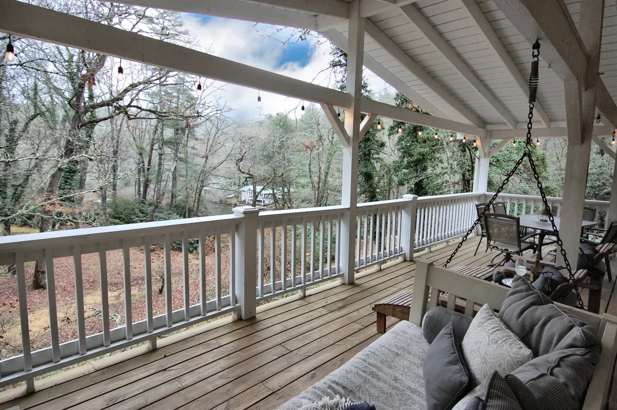 A spacious wooden deck nestled among mature trees, featuring a hanging swing and a dedicated outdoor seating area. The deck overlooks natural scenery with partial views of the lake, and the setup invites relaxation, conversation, and connection with nature.