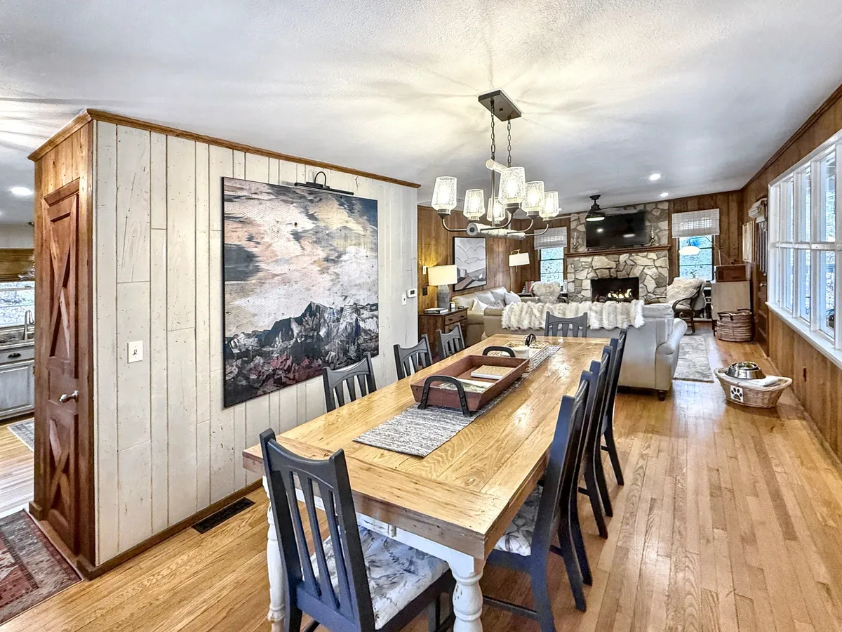 A dining area with a spacious table, elegant light fixtures, and a fireplace in the background, accented by artwork on the walls. The setting is ready for shared meals or game nights with friends and family.