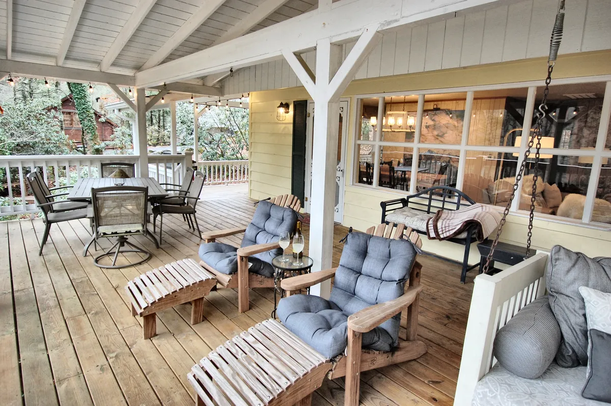 An inviting covered porch setup with wooden chairs, a dining table, and a swing bench adorned with decorative pillows. Soft natural light enhances the cozy, comfortable vibe, with hints of surrounding trees and nature in view.
