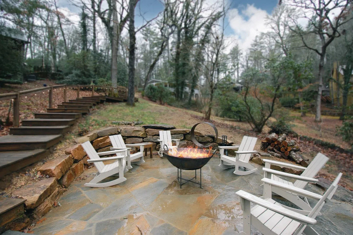 A cozy outdoor stone fire pit patio is surrounded by Adirondack chairs, creating an inviting space for guests to gather around the fire. The area is nestled in nature, with greenery and trees providing a scenic backdrop, and the arrangement hints at evenings spent sharing stories and roasting s'mores.