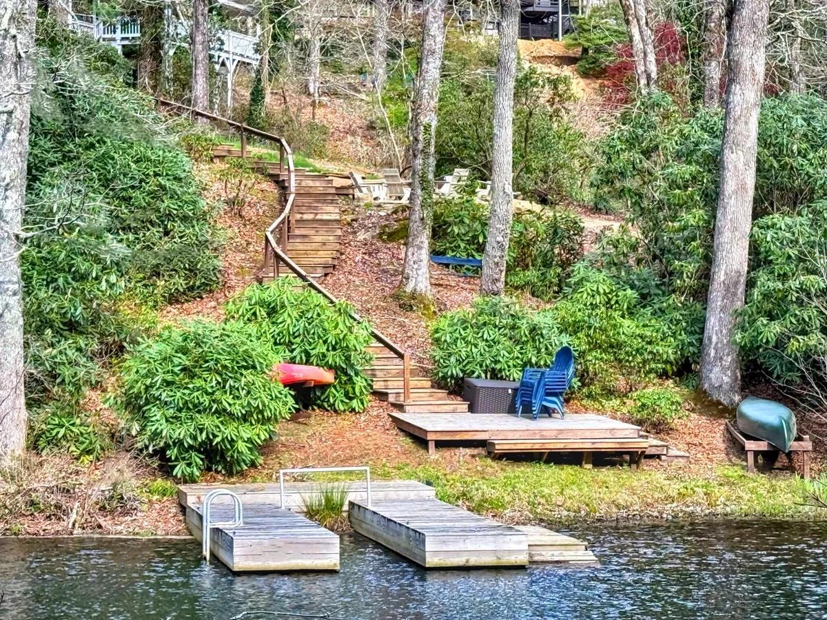 The image showcases the tranquil lakefront exterior of the home, with wooden steps leading down to a private dock surrounded by lush green foliage. Comfortable outdoor seating invites guests to relax by the water, while glimpses of kayaks suggest adventure and fun. The serene setting is highlighted by the possibility of sunset views over the lake, creating an inviting and peaceful atmosphere.
